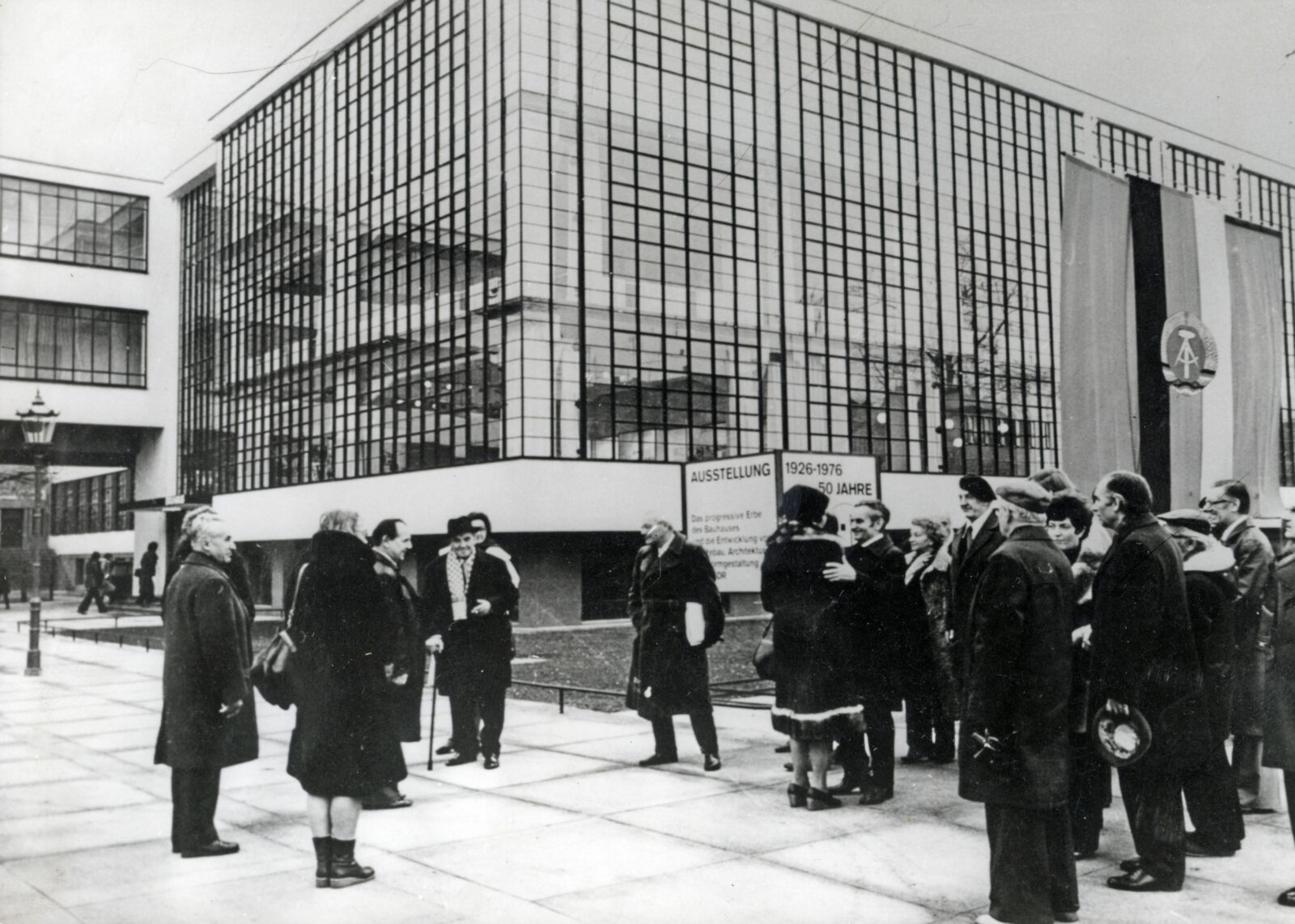 Viele Menschen stehen vor der Ecke des Bauhausgebäudes – einige begrüßen sich freudig. Im Hintergrund weht die Fahne der Deutschen Demokratischen Republik.
