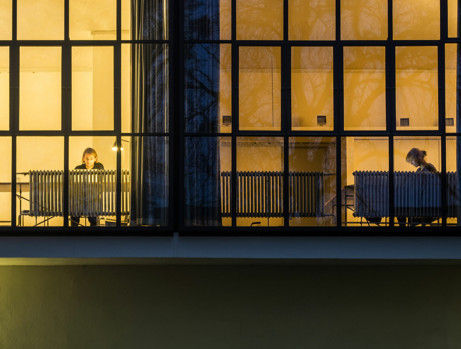 Blick in die hell erleuchteten Atelierfenster des Meisterhaus Muche-Schlemmer. An dem Schreibtischen sitzt ein junger Mann auf der linken Seite und eine junge Frau auf der rechten Seite des Doppelhauses.