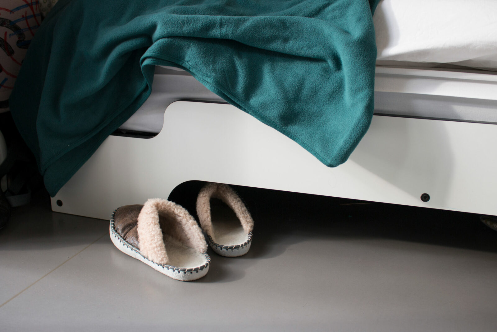 Detailaufnahme von zwei Pantoffeln, die vor einem Bett stehen. Eine türkise Zudecke hängt aus dem Bett heraus.