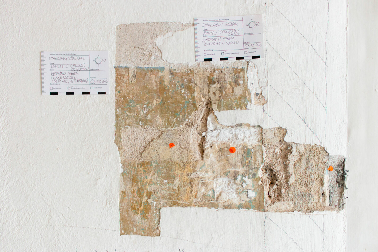 Ein Stück Wand zeigt die verschiedenen Farbschichten. Daneben kleben Kleine Zettel, auf denen handschriftlich genauere Angaben zur Farbschicht gemacht werden.