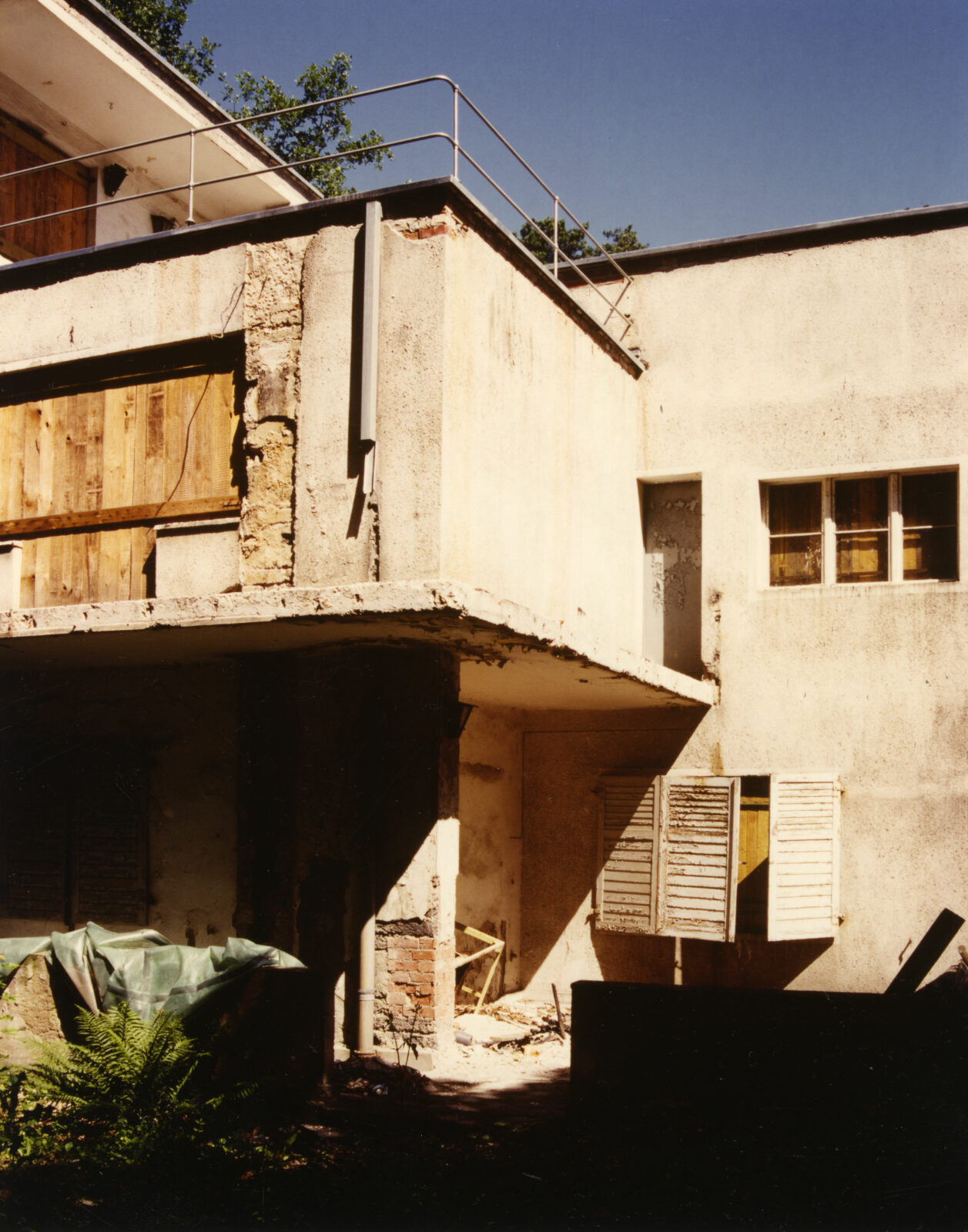 Detail einer Hausecke im desolaten Zustand. Der Balkon ist verfallen, die Fensterläden hängen schief. Die Fensteröffnungen sind mit Holzbrettern verschlossen.