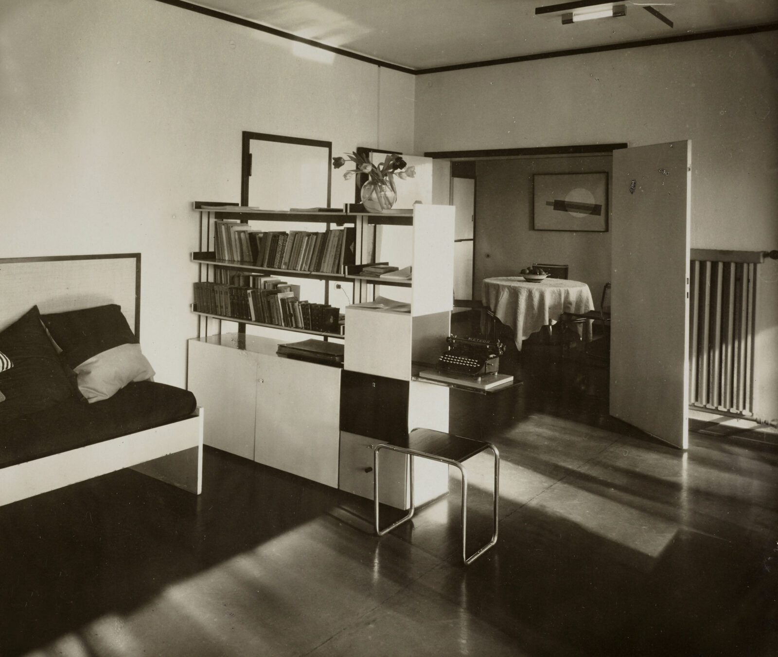 Ein modernes Bücherregal mit extra Ausschub für eine Schreibmaschine ragt in das Wohnzimmer des Meisterhauses Mohly hinein. Daneben steht ein Stahlrohrhocker. Im Hintergrund die geöffnete Tür zum Esszimmer. Auf dem Tisch steht eine Obstschale. An der Wand hängt eine Grafik.