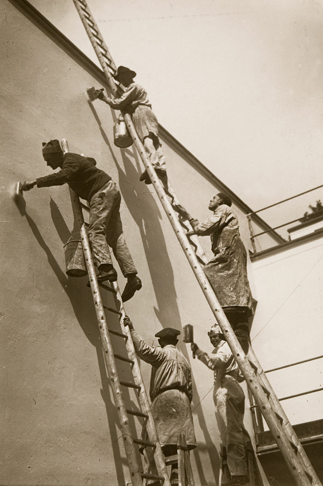 Männer mit Pinseln und Farbeimern in der Hand stehen auf langen Leitern, die an die Wand des Meisterhauses gelehnt stehen.