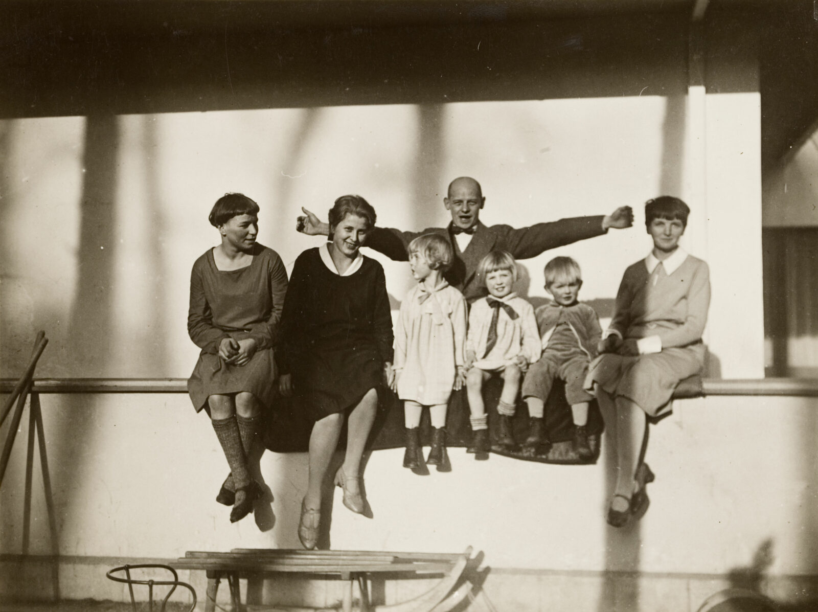 Der Bauhaus Meister Oskar Schlemmer steht in einer freudig ausladenden Geste mit ausgebreiteten Armen hinter eine Gruppe von drei Frauen und drei Kindern, die auf der Mauer der Terrasse des Meisterhauses sitzen.