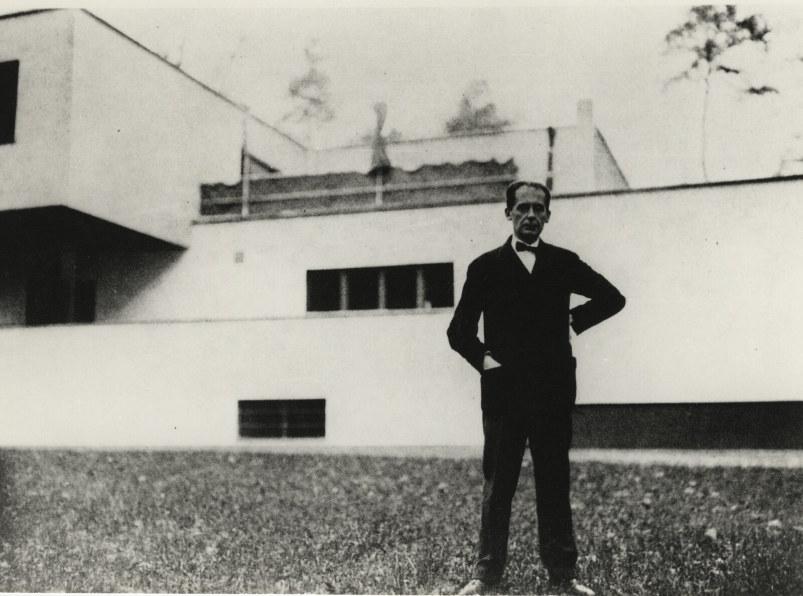 Walter Gropius steht vor seinem Wohnhaus. Einen Arm hat er in die Hüfte gestemmt – die andere Hand ist in der Jacketjacke. Er trägt eine Fliege.
