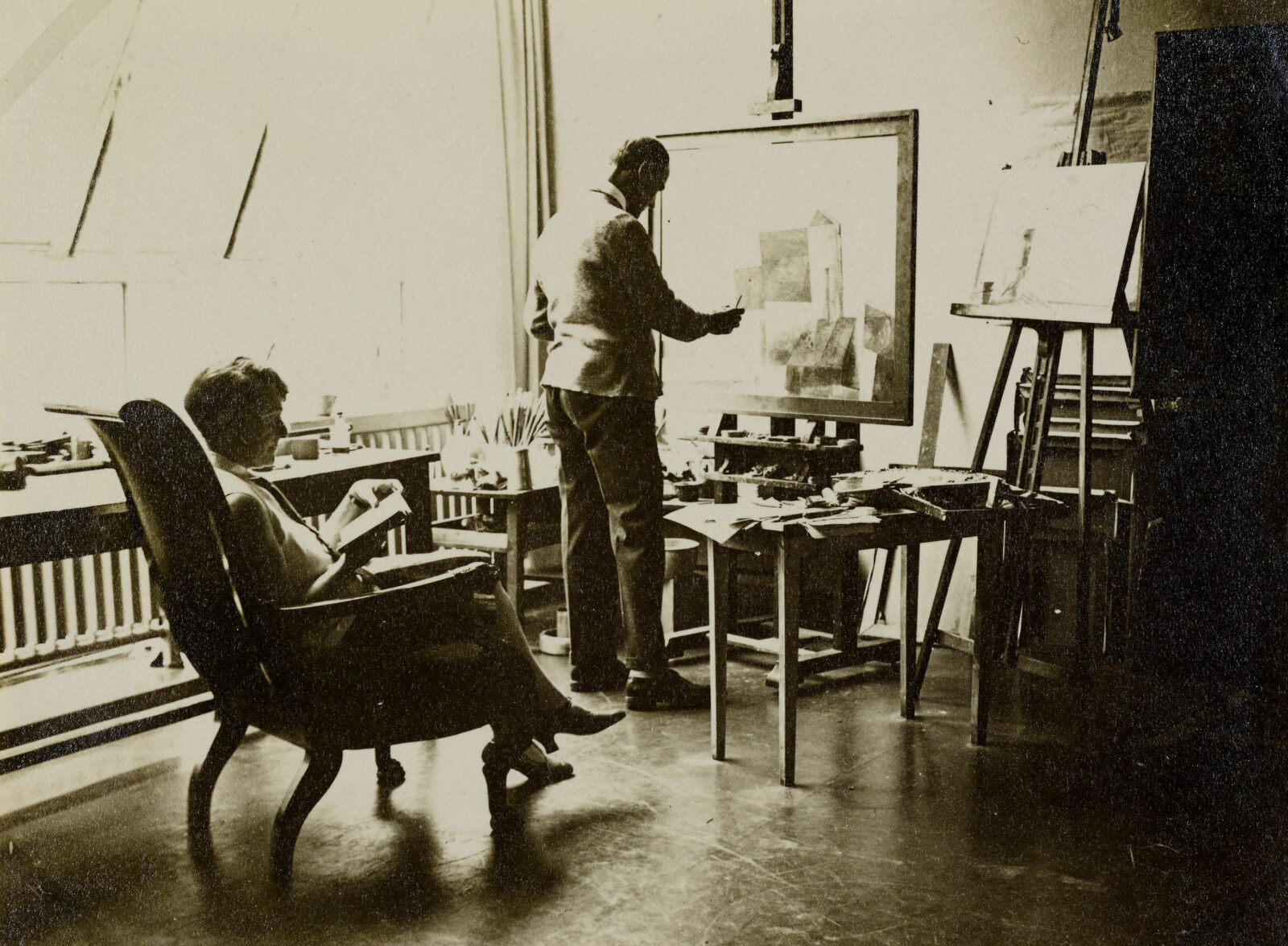 Der Bauhaus Meister Lyonel Feininger steht an einer Staffelei im Atelier und malt. Seine Frau sitzt daneben in einem Sessel und liest in einem Buch.