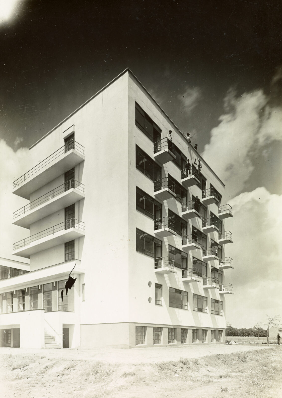 Historisches Foto vom Atelierhaus mit Balkonen von 1927, in schwarz-weiß. Auf einzelnen Balkonen im 3. und 4. Obergeschoss sowie auf dem Dach des Gebäudes stehen Bauhäusler und winken.