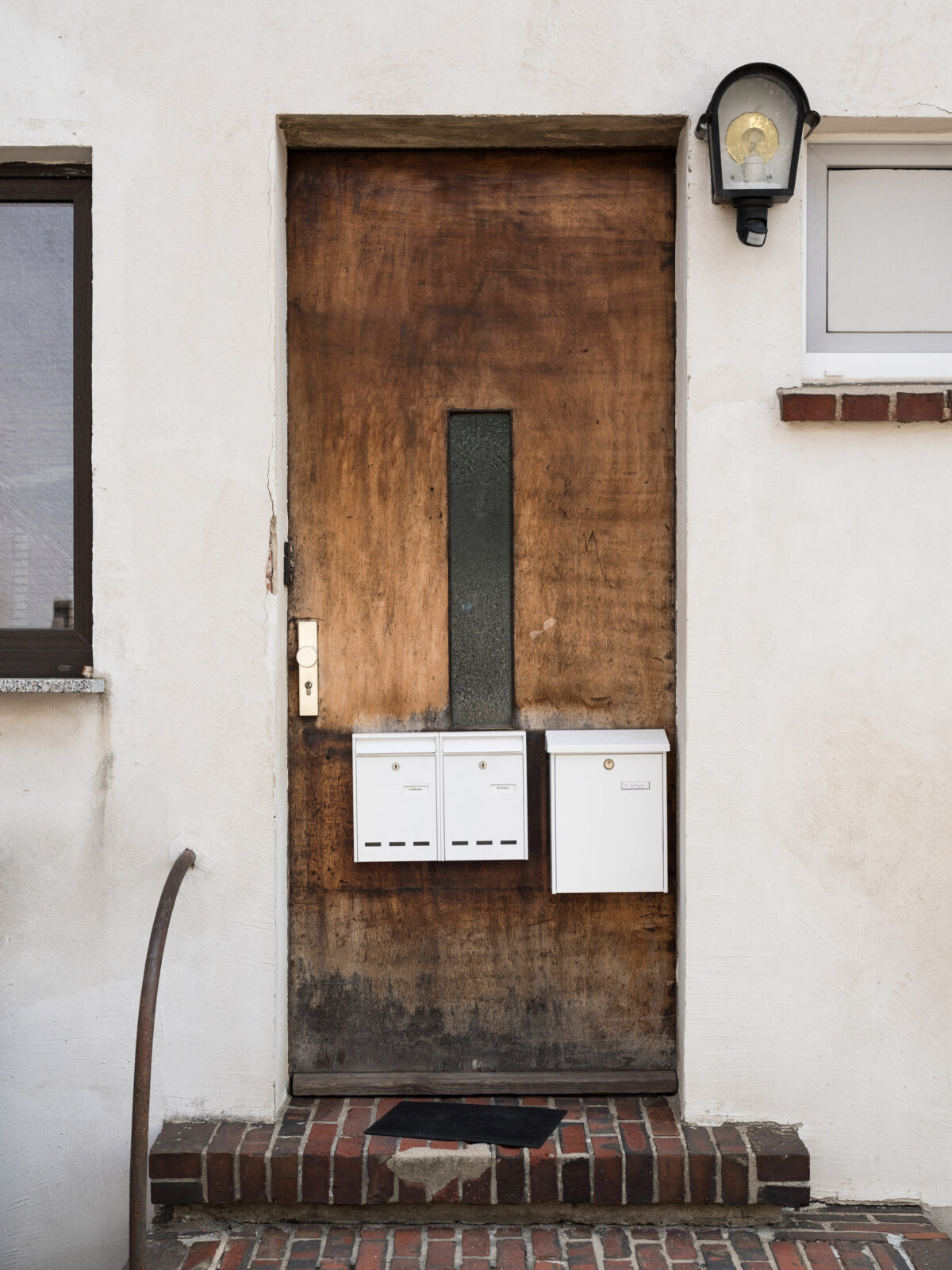 Hölzerne Eingangstür mit drei Briefkästen – davor zwei Stufen aus Klinkersteinen und gebogenem rundem Handlauf.