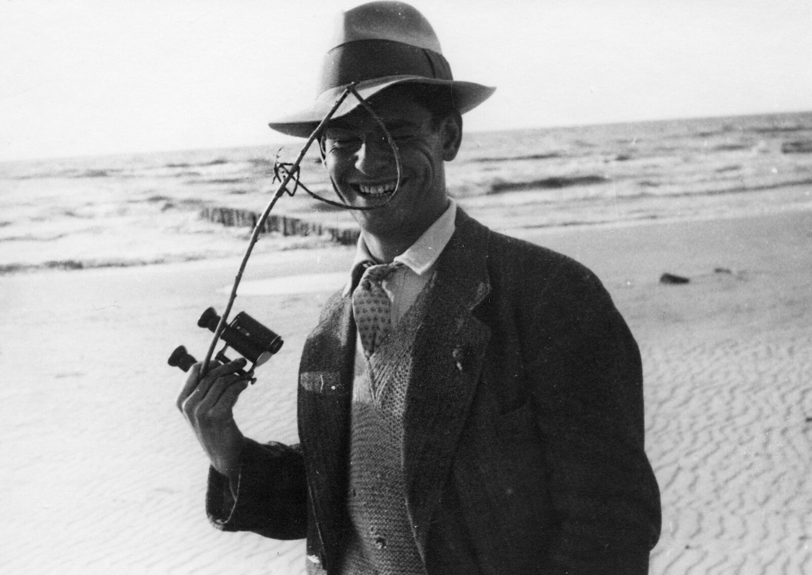T. Lux Feininger mit Hut am Meer. In der Hand hält er ein Fernglas und einen gebogenen Stock, durch den er lächelnd dem Betarchter entgegenschaut.